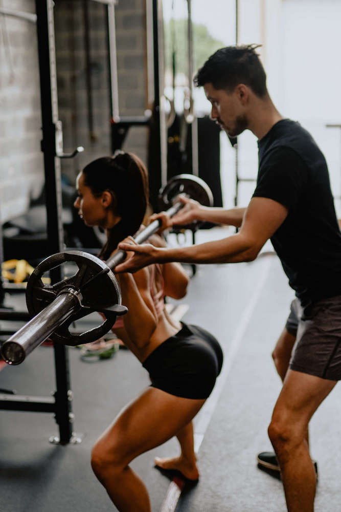 Woman doing barbell squats in front of a mirror with a personal trainer